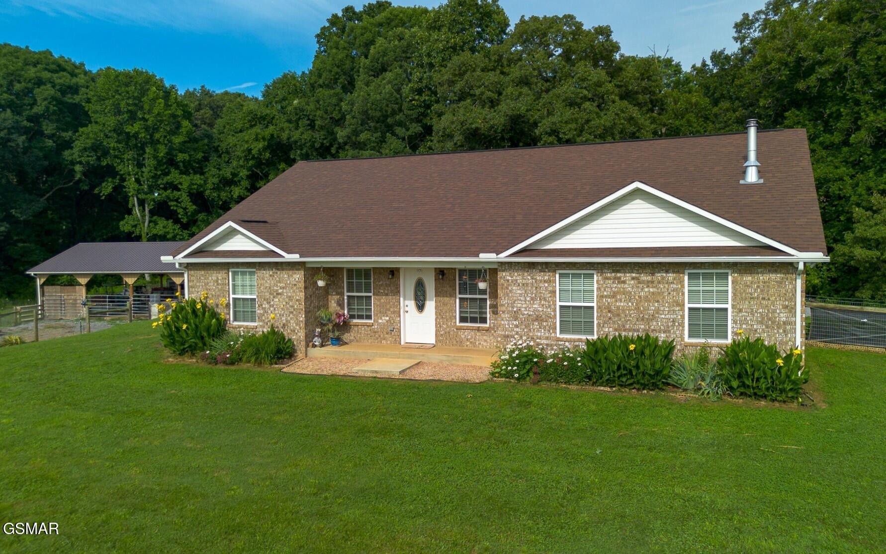 Brick ranch home with a broad lawn, attached carport, and wooded backdrop.