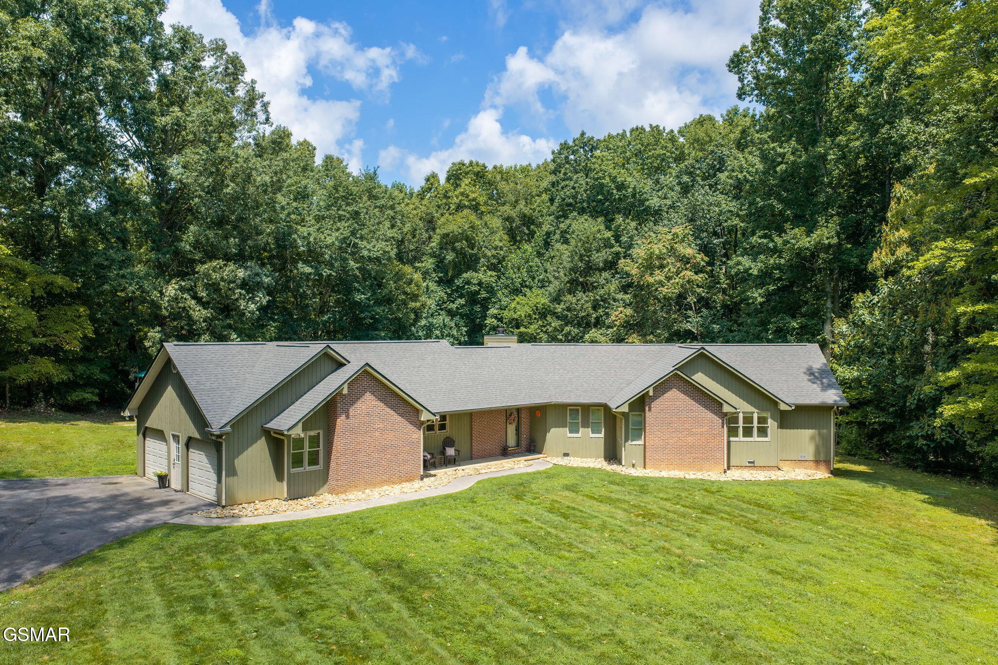 Single-story green and brick home with two garage doors, a broad lawn, and woods behind it.
