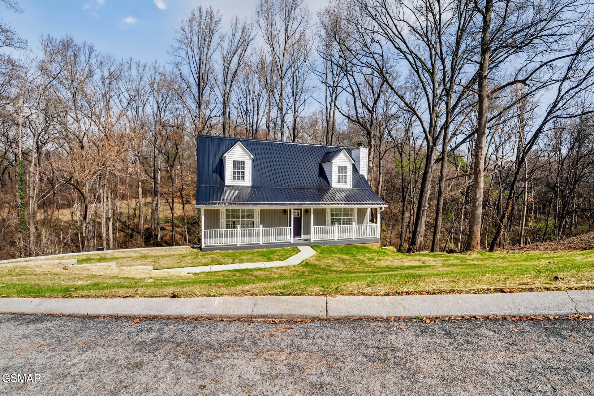 White house with a dark metal roof, two dormers, and a covered porch among leafless trees.