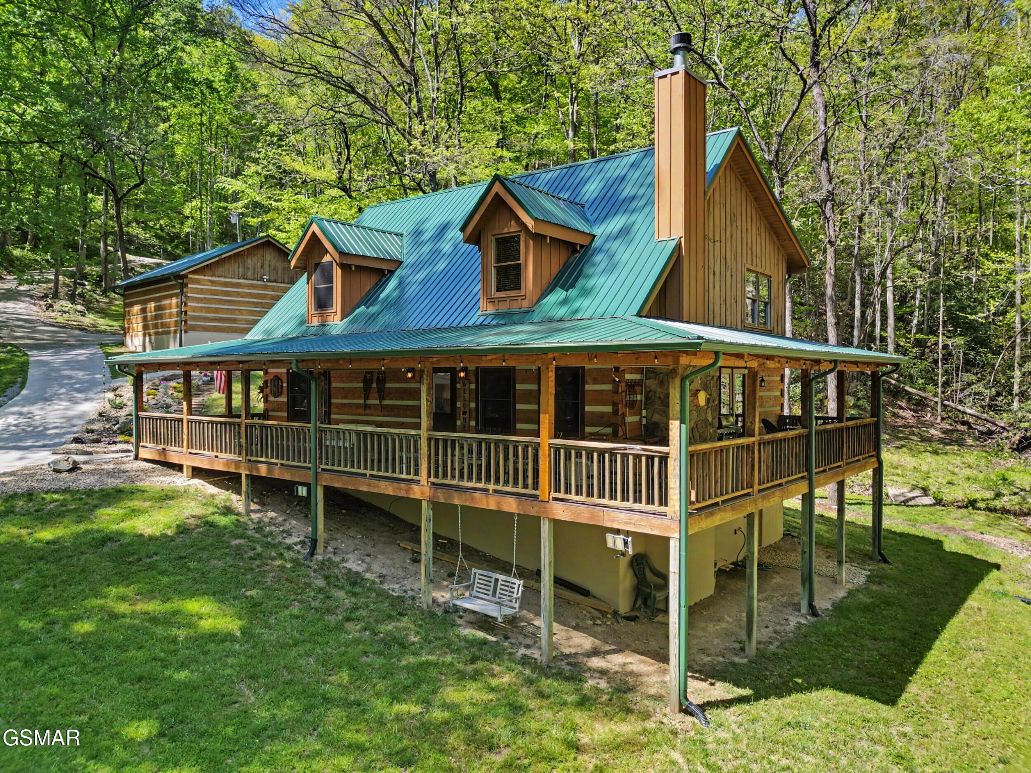 Wood-sided cabin with a green metal roof and wraparound porch among leafy trees.