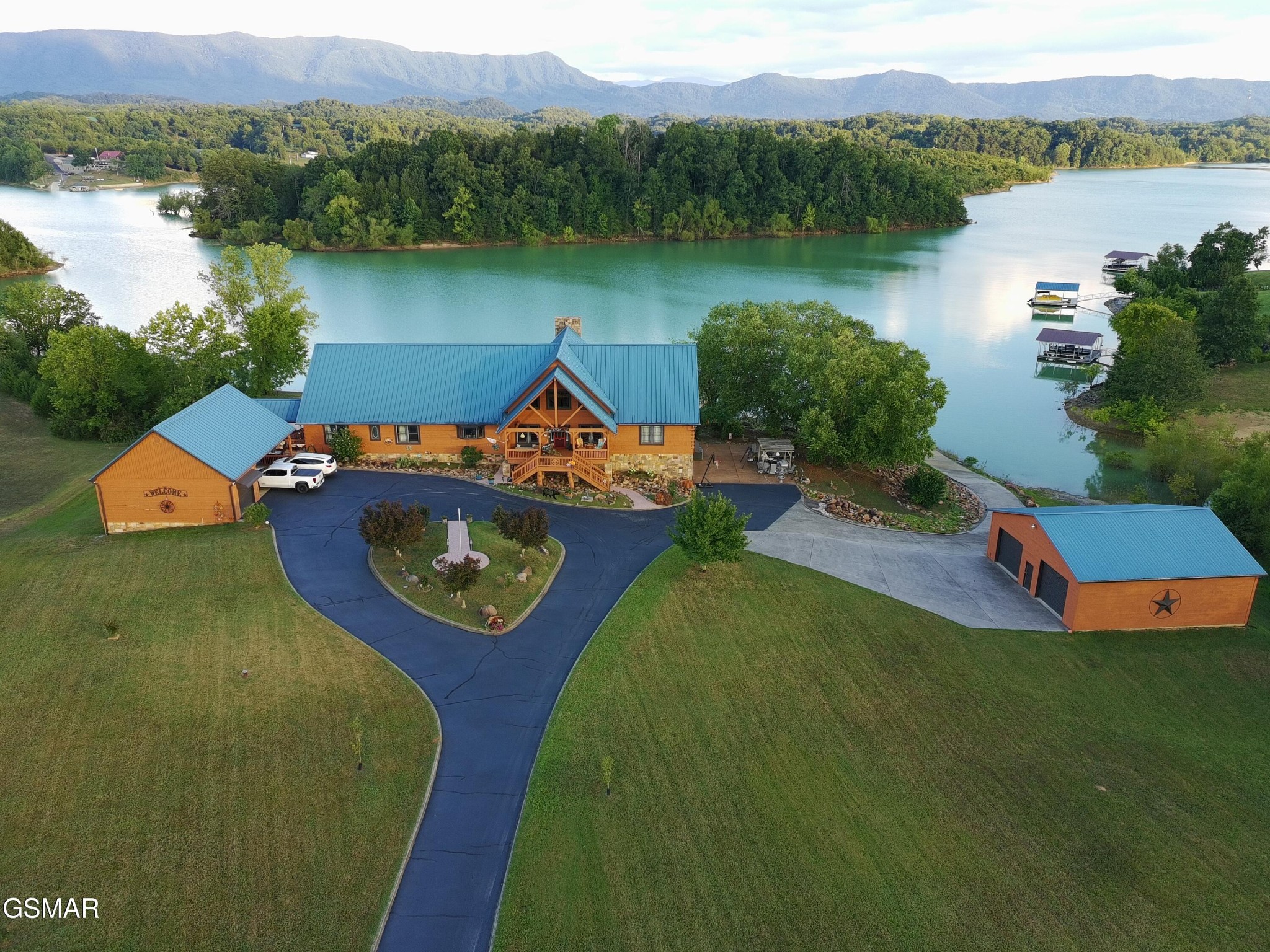 Aerial view of a blue-roofed lakeside home with a circular driveway, wooded island, and distant mountains.