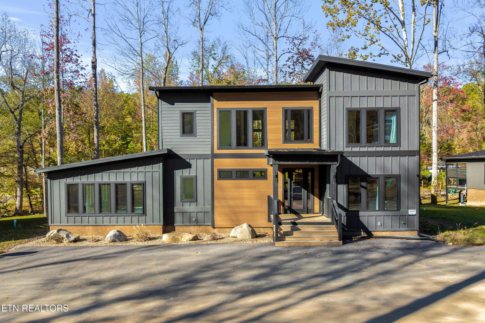 Modern two-story home with charcoal siding, wood accents, and a covered entry among autumn trees.