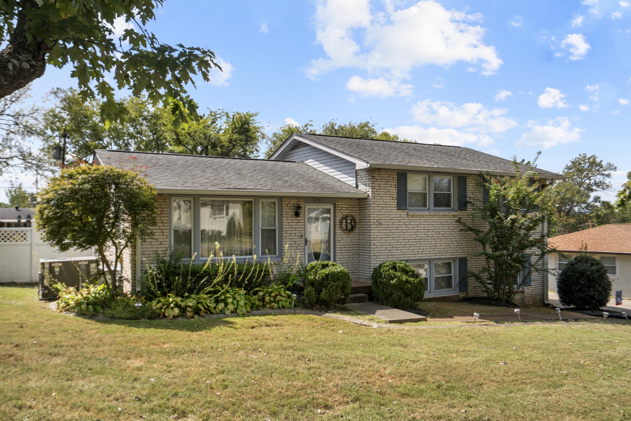 Light brick two-level home with dark shutters, front landscaping, and a broad lawn.