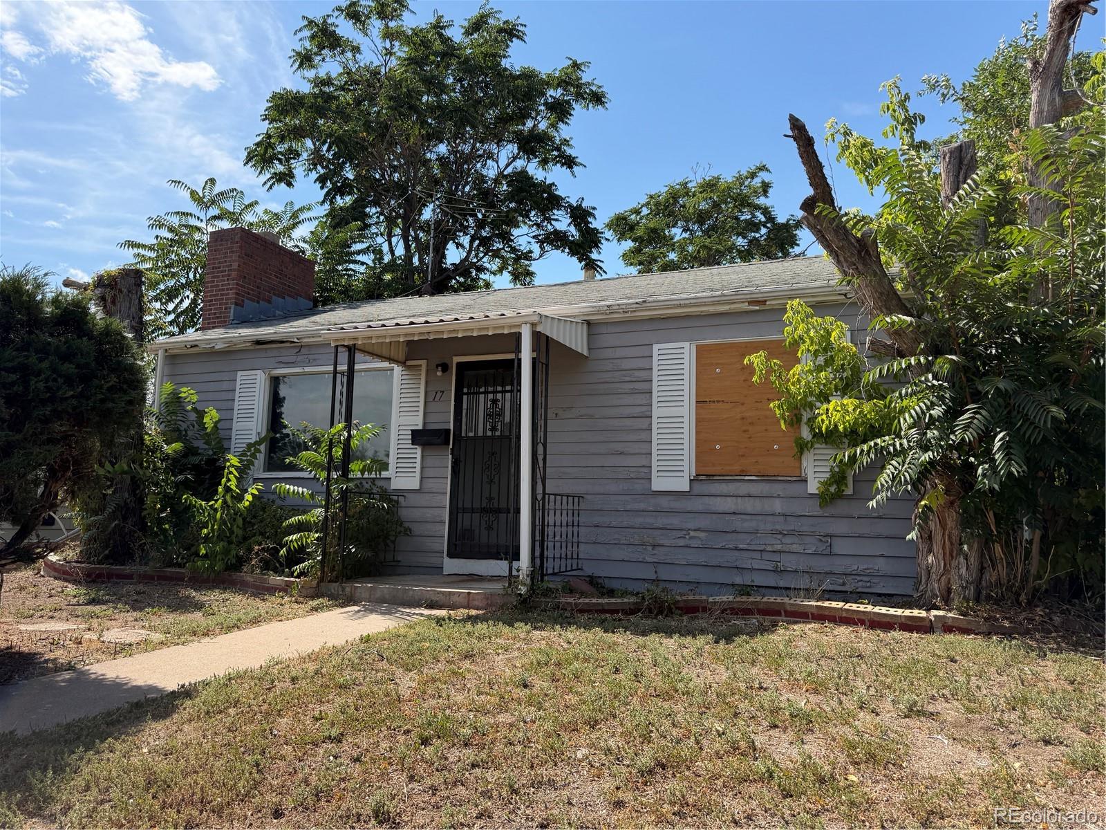 Gray single-story house with a brick chimney, barred entry, and a plywood-covered window.