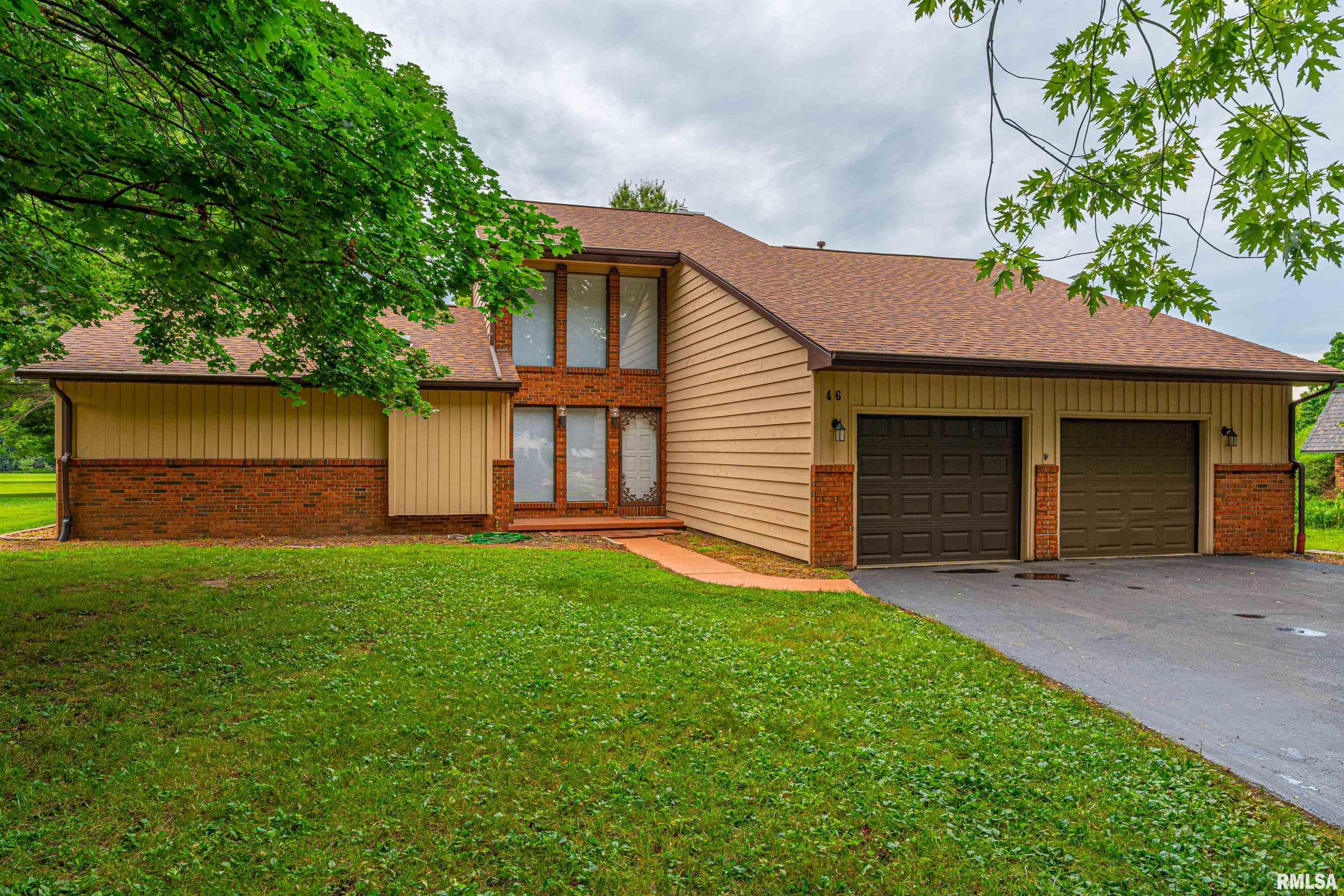 Two-story tan-and-brick house with an attached two-car garage, framed by leafy trees and a broad lawn.
