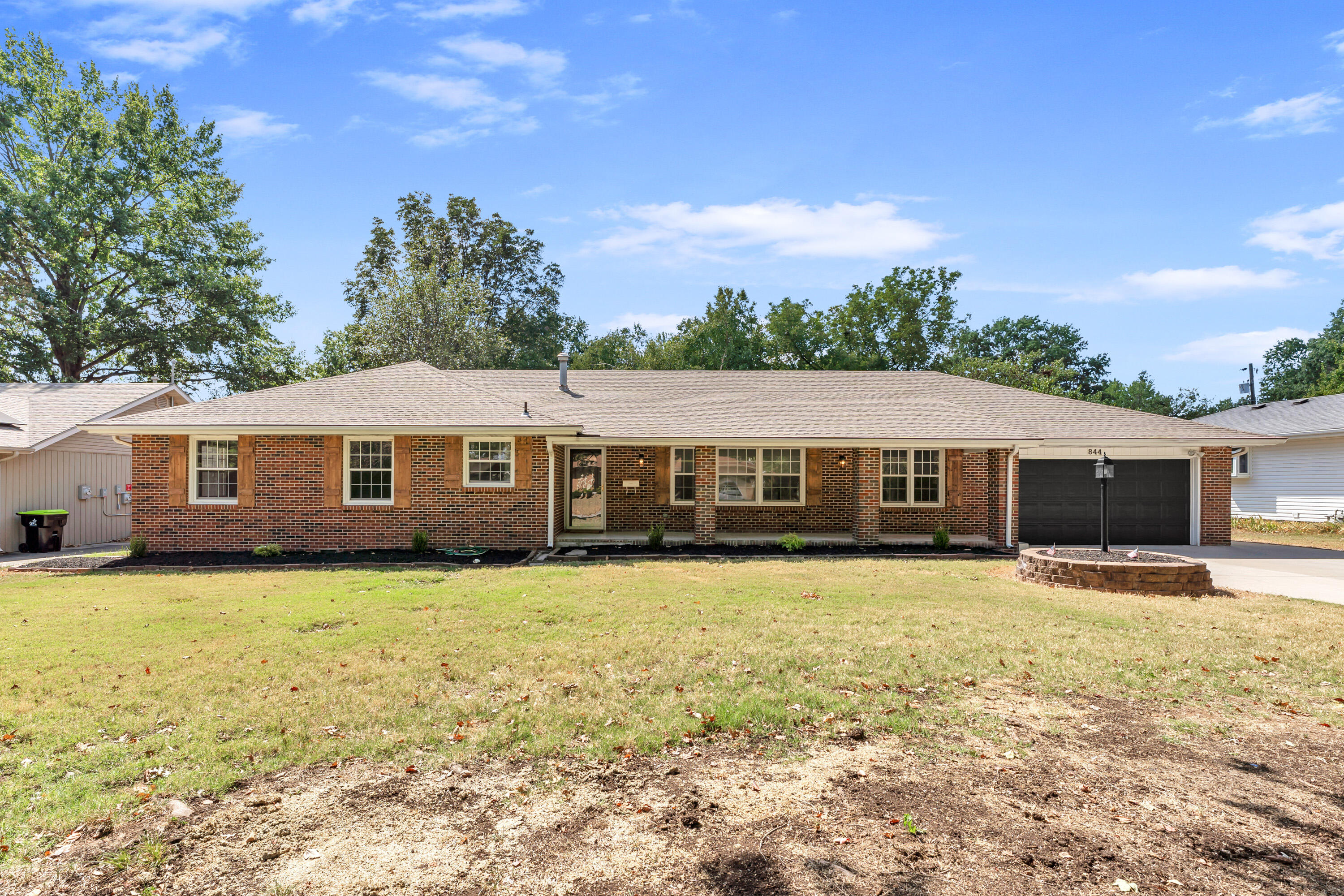 Single-story brick ranch with an attached black garage, broad lawn, and mature trees.