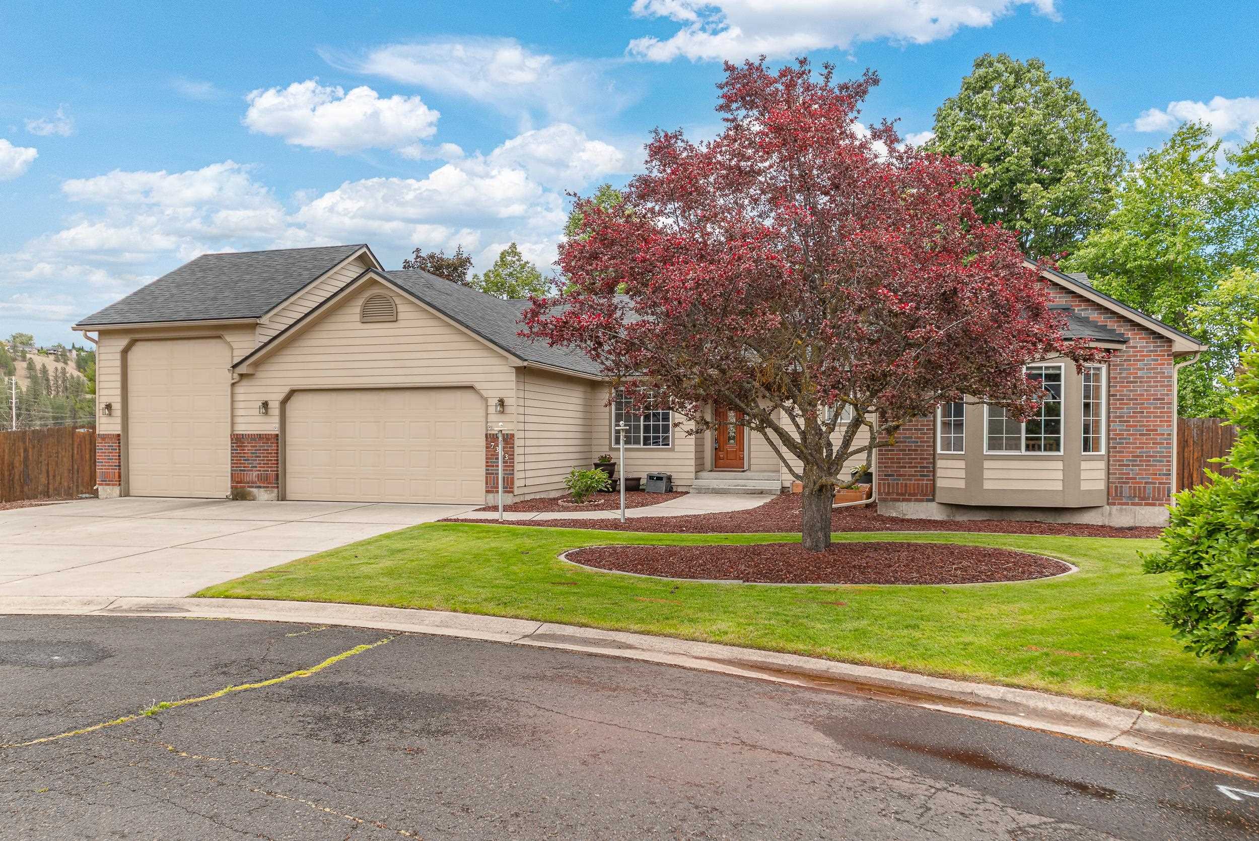 Beige home with brick accents and a two-car garage behind a large red-leaf tree.