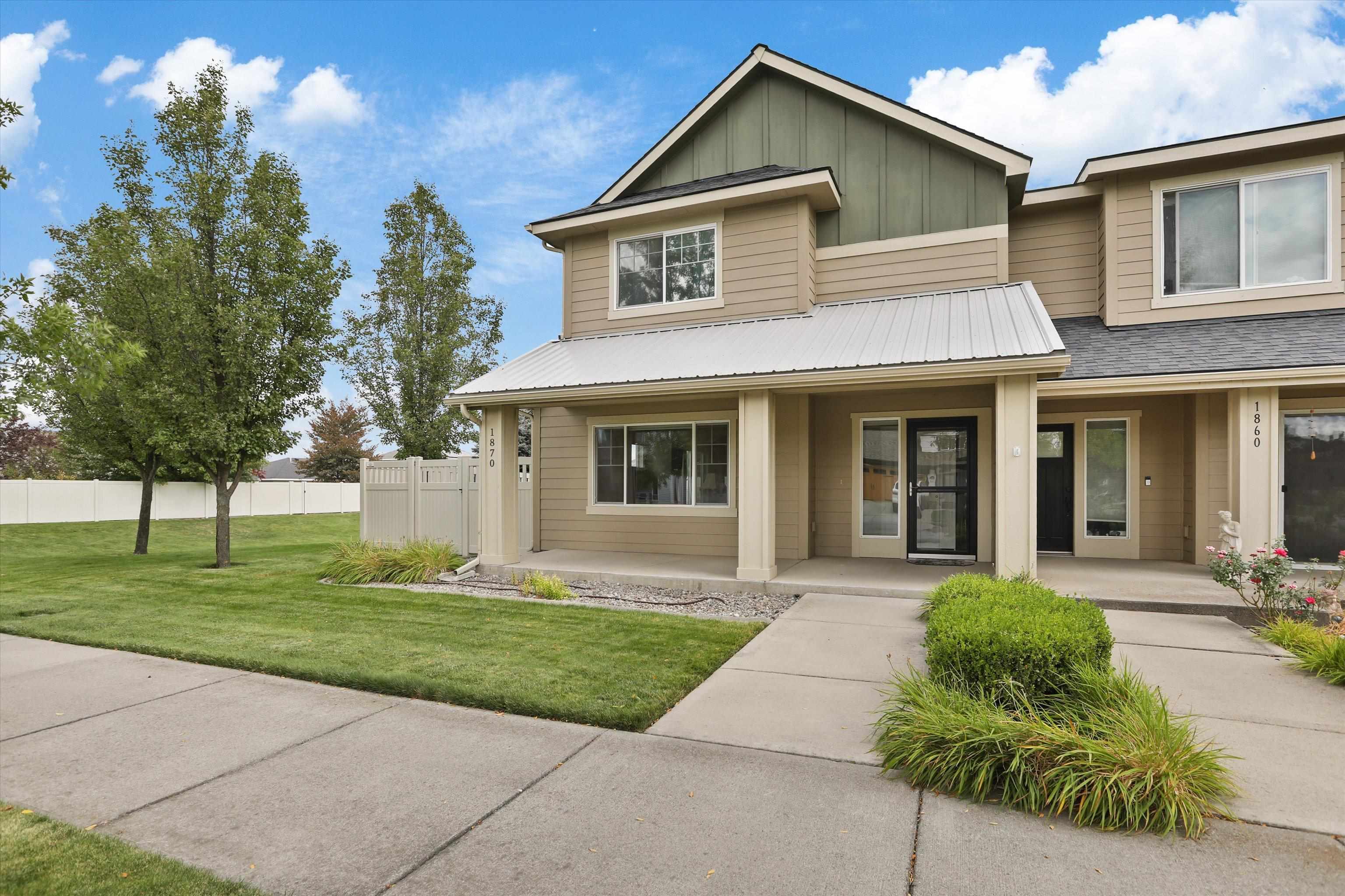 Attached beige townhomes with green gables, covered entries, landscaped lawns, and a concrete walkway.